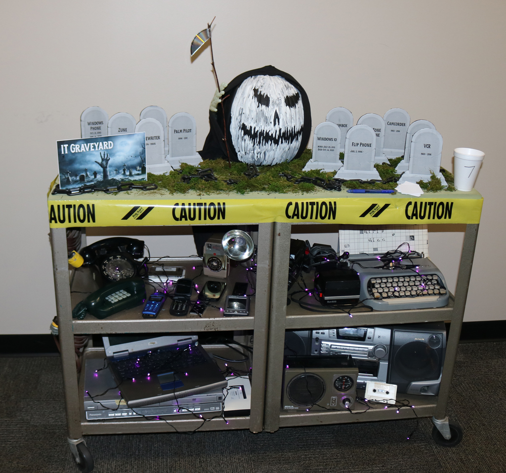 IT Graveyard - cart of outdated electronics, a grave yard with grave stones of out dated devices, and a black grim reaper pumpkinand with a white face