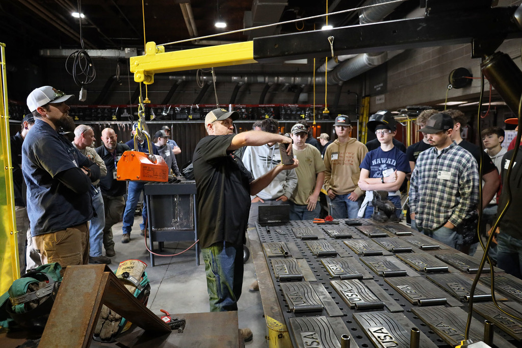 Weldng contest students gathered around  while a metal plate with USA cut into it and wavey lines like a flag next to it an below