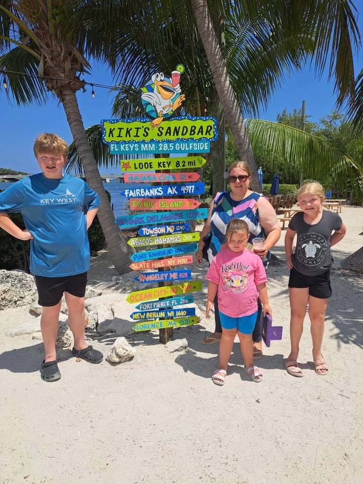 Mrs Mueller and her kids by a sign with directions arrows with names and distances to different destinations including Fiarbanks Alaska