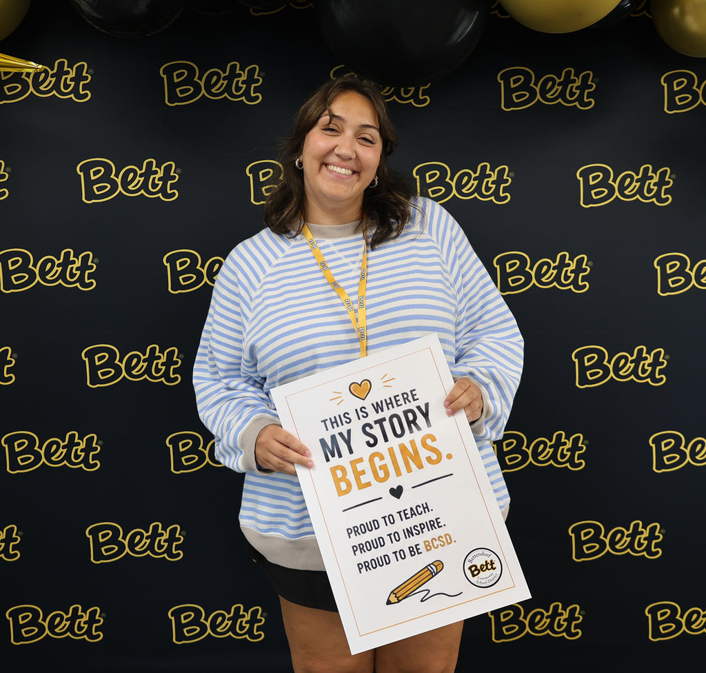 Gabby Baldry with a sign: Heart. This is were my story begins. Proud to teach. Proud to inspire. Proud to be at BCSD. Pencil. Bettendorf Community School District logo with Bett graphic