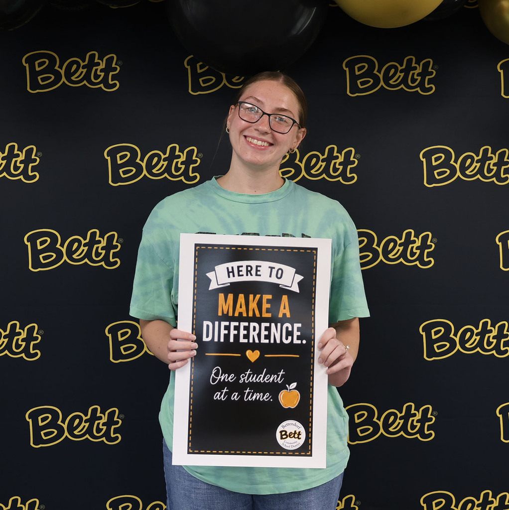 Olivia Sholl BMS holding a sign: Here to make a difference. Heart. One student at a time. Apple. Bettendorf Community School District logo with Bett graphic.