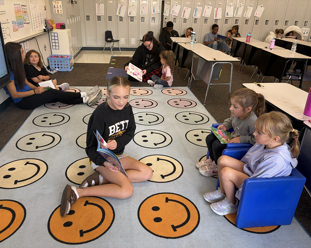 A HS student reading to two student on a carpet of smiley faces