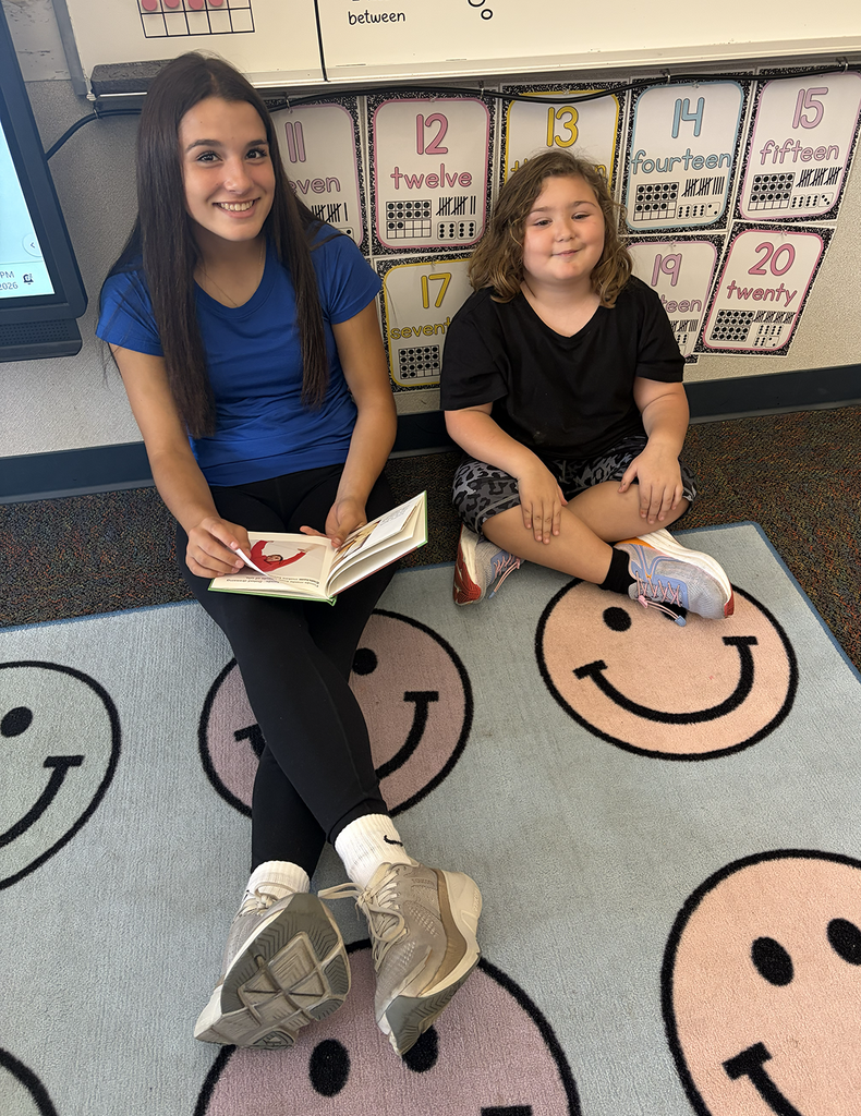 A HS and NA student sitting on the floor with a book smiling.