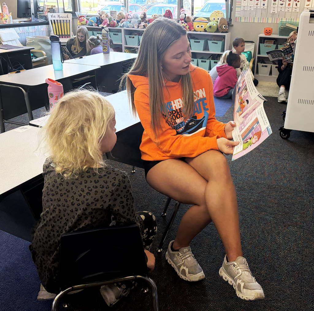 A HS student reading to a NA student in a classroom