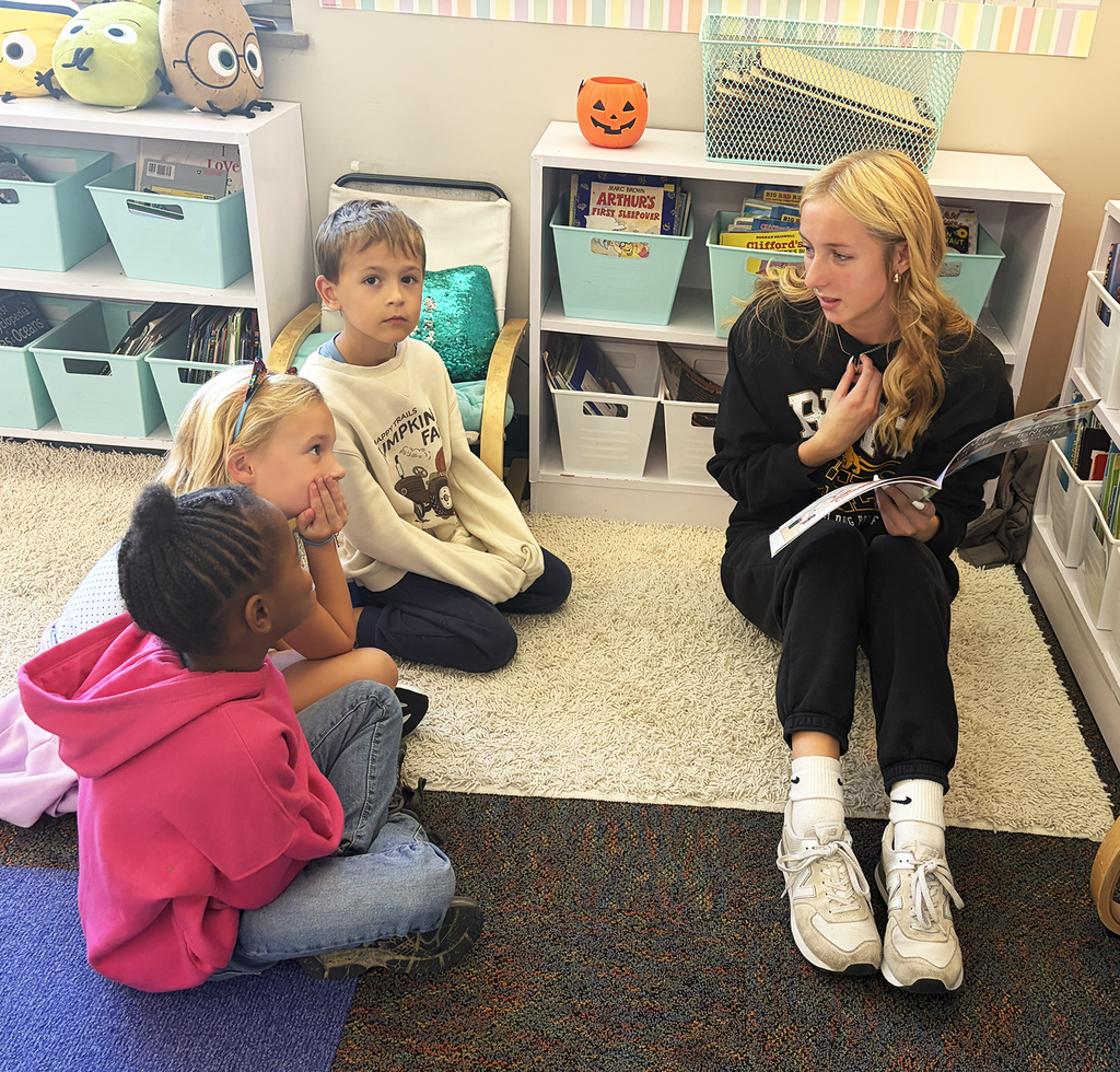 A HS student reading to a group of three students in a classroom