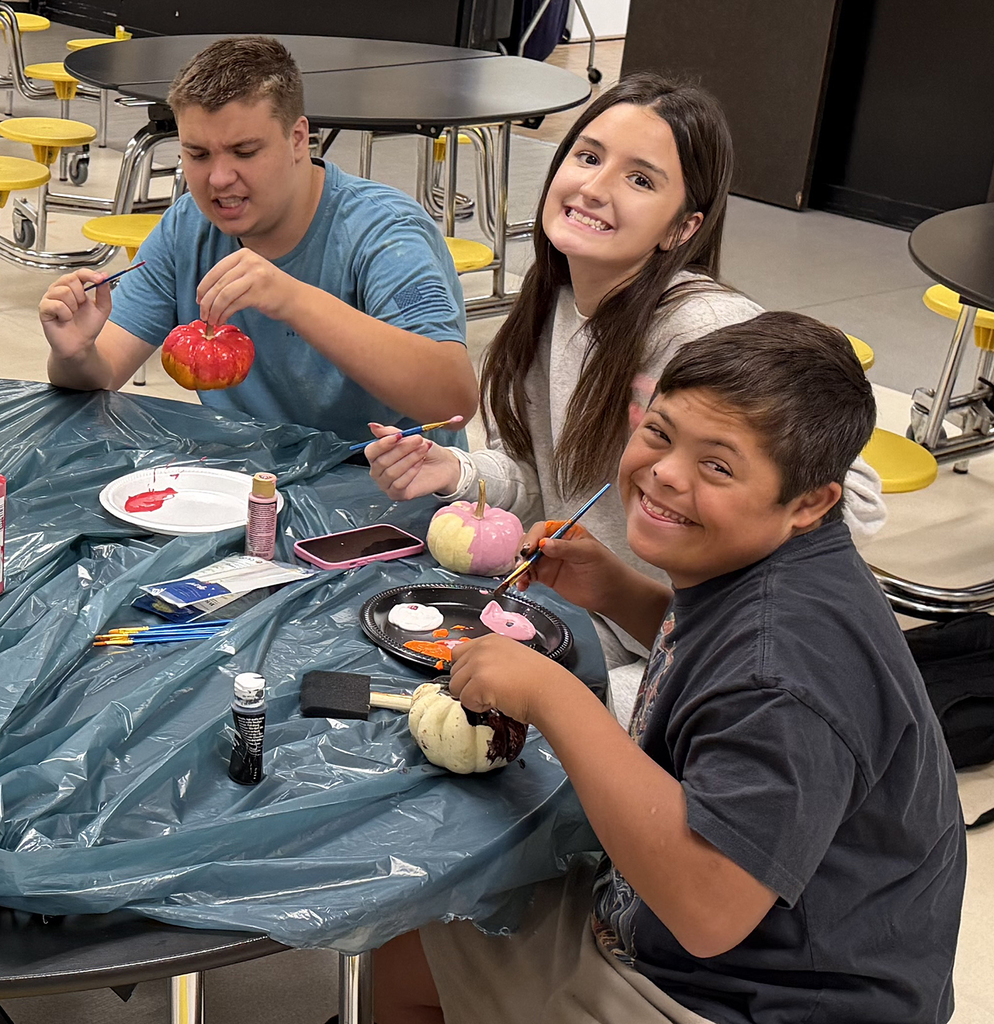 Two students smiling at the camera while working on their pumpkin. A third student busy painting.