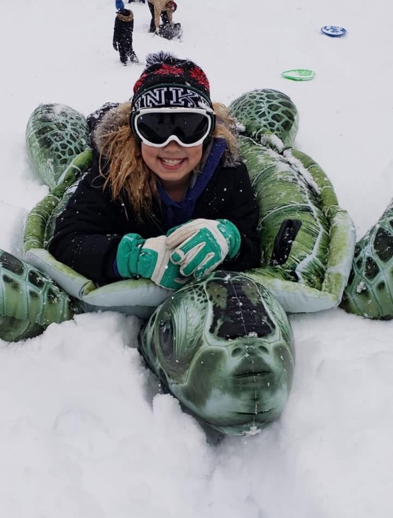student smiles for camera while laying on turtle sled in snow