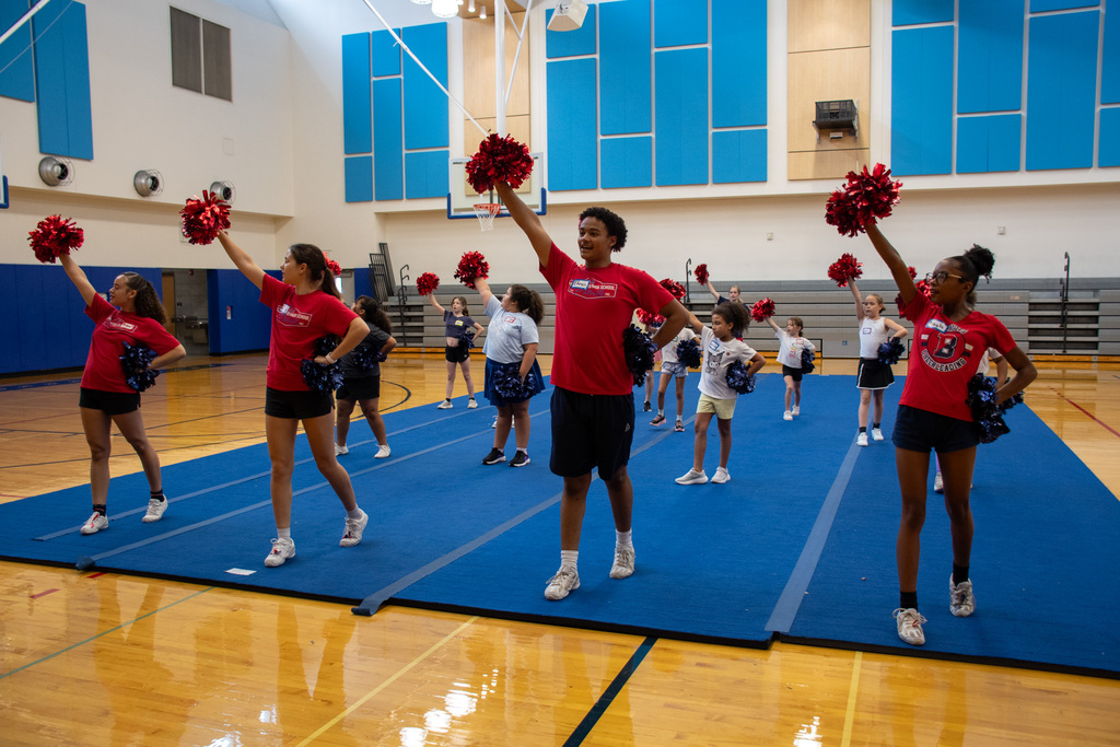 varsity cheerleaders lead younger students in cheer with everyone raising a pom pom above their head