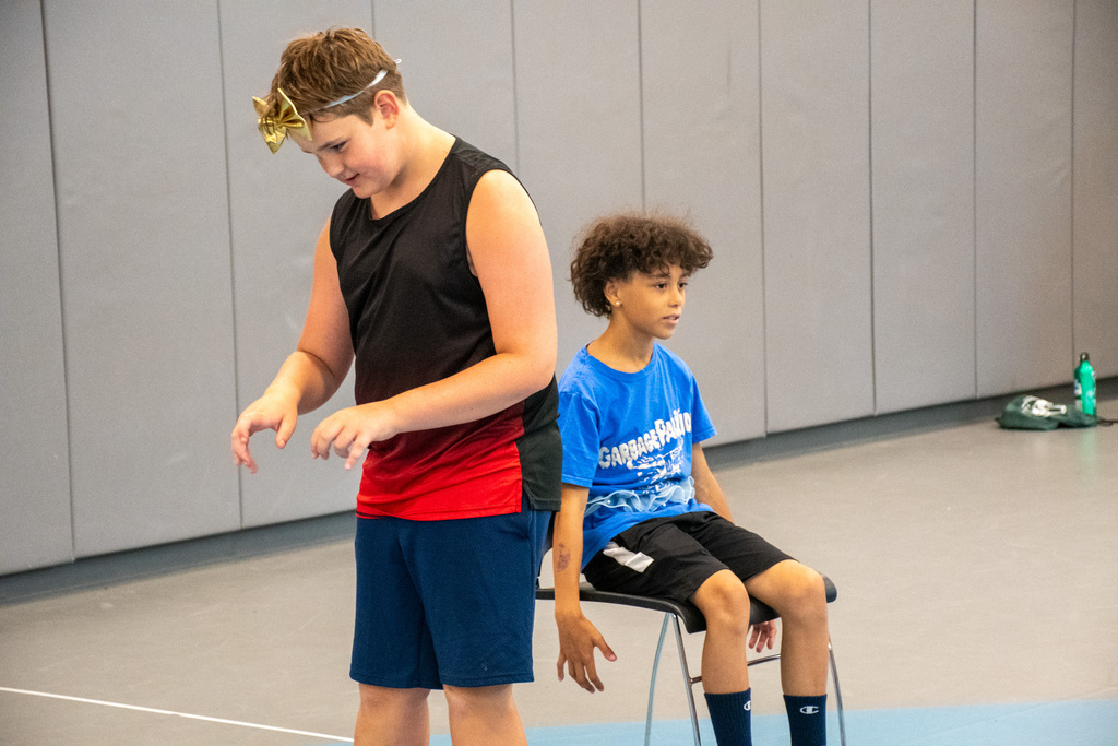 student wearing oversized bowtie around his head pretends to type on a computer while second student sits in chair behind him during theatre workshop