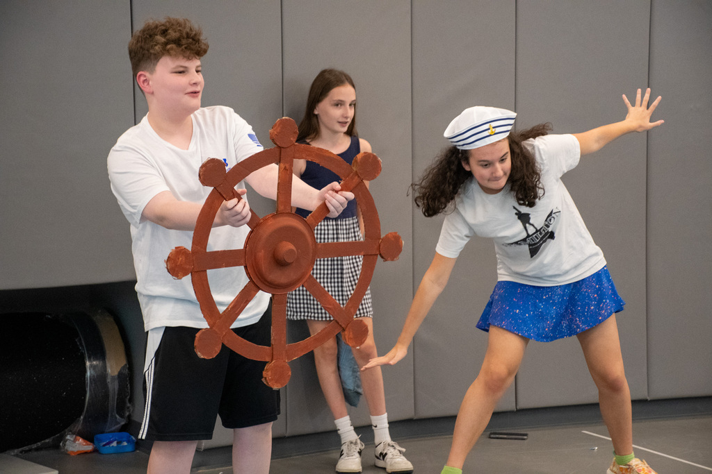 student holds large ship steering wheel while second student pretends to keep balance and a third stands against the wall during skit at theater workshop