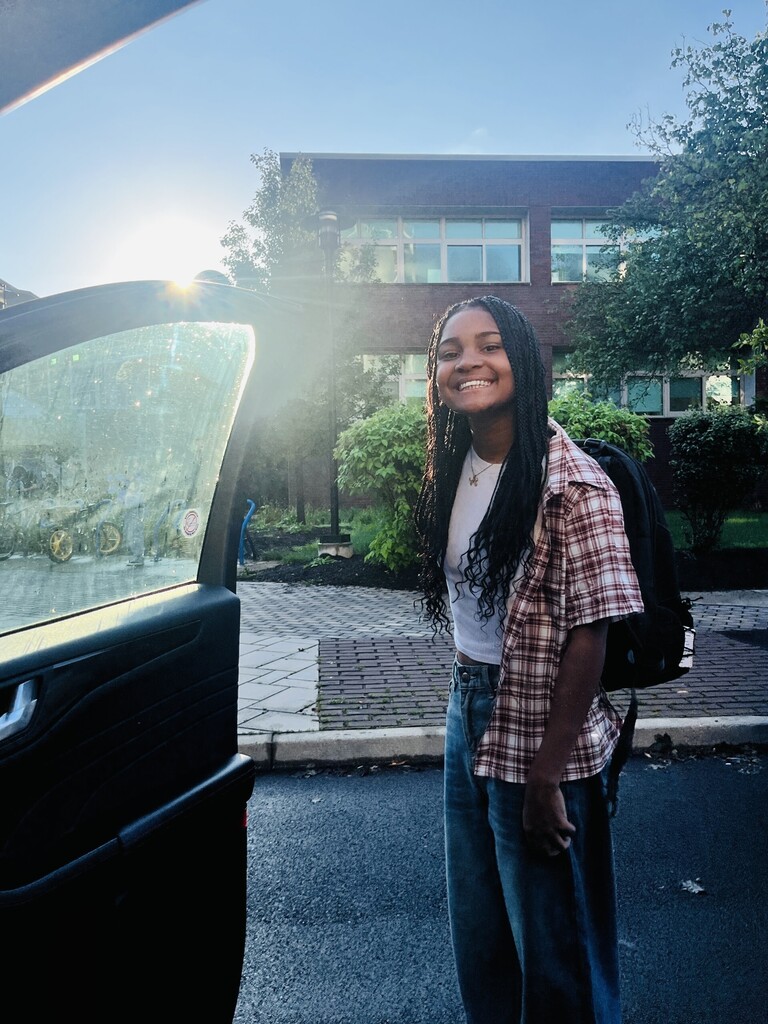student smiles for camera after getting out of car with school in background