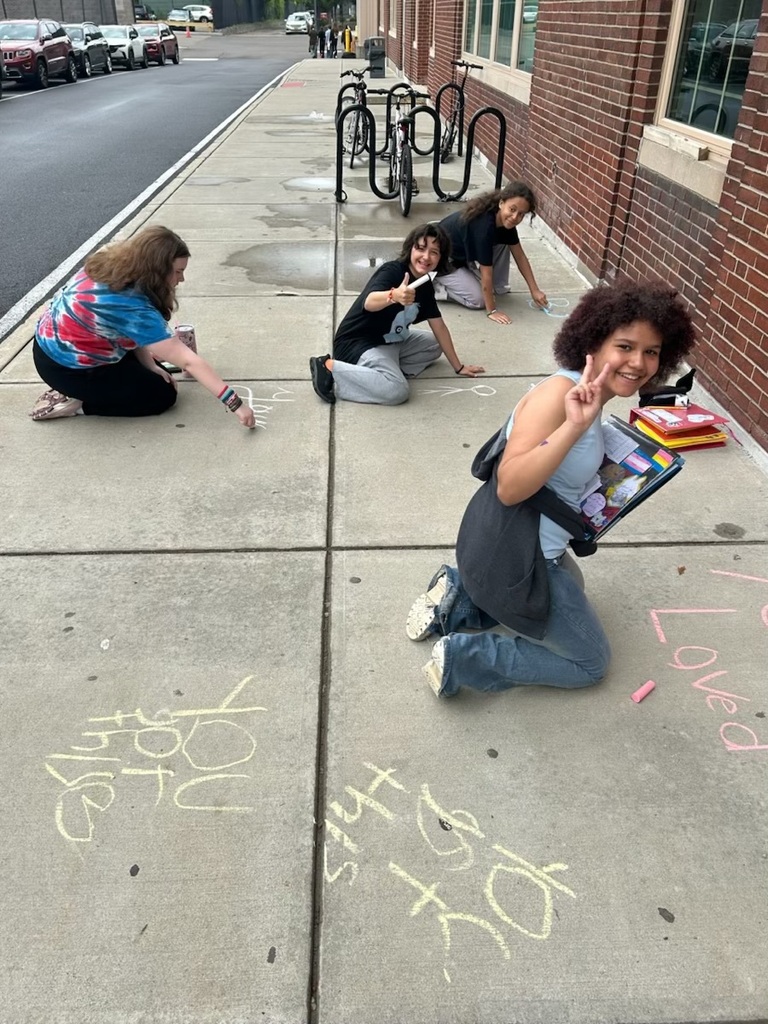 east middle students smile for camera while writing positive chalk messages outside their school