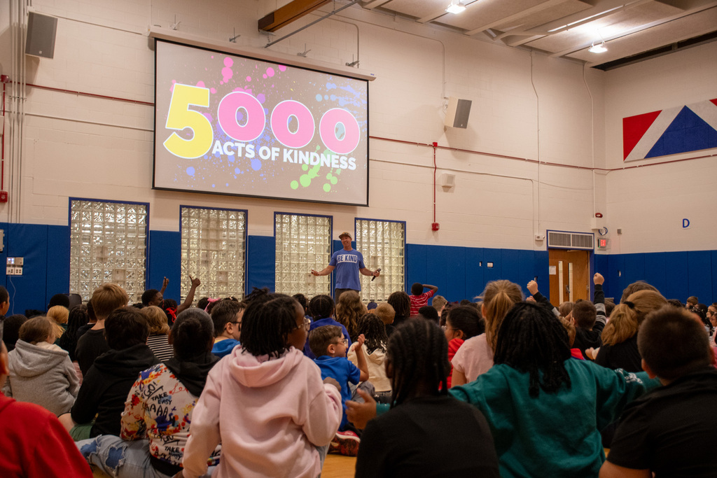 screen that says "5000 acts of kindness" in gymnasium with students seated on floor looking on