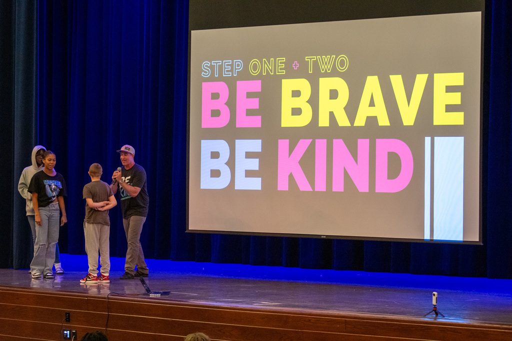 east middle school students on stage with "Think Kindness" presenter Brian Williams; graphic that says "step one be brave, step two be kind" is on screen in background