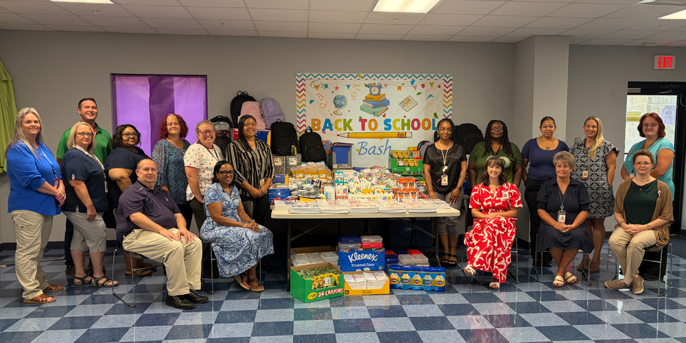 A group of adults stand together smiling around collected school supplies.