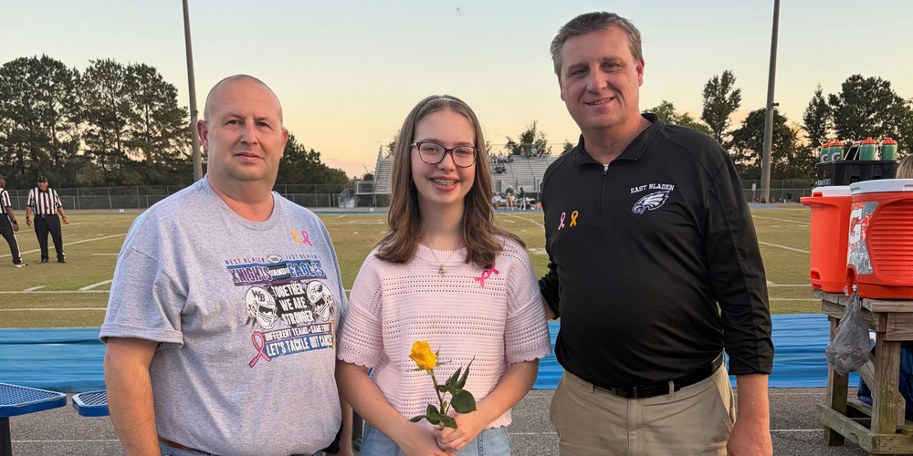 A high school student wearing a pink shirt and holding a yellow rose stands smiling between two administrators infront of the football field.