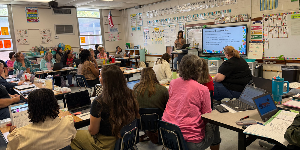 A group of educators sit at tables in an elementary classroom listening to a teacher.