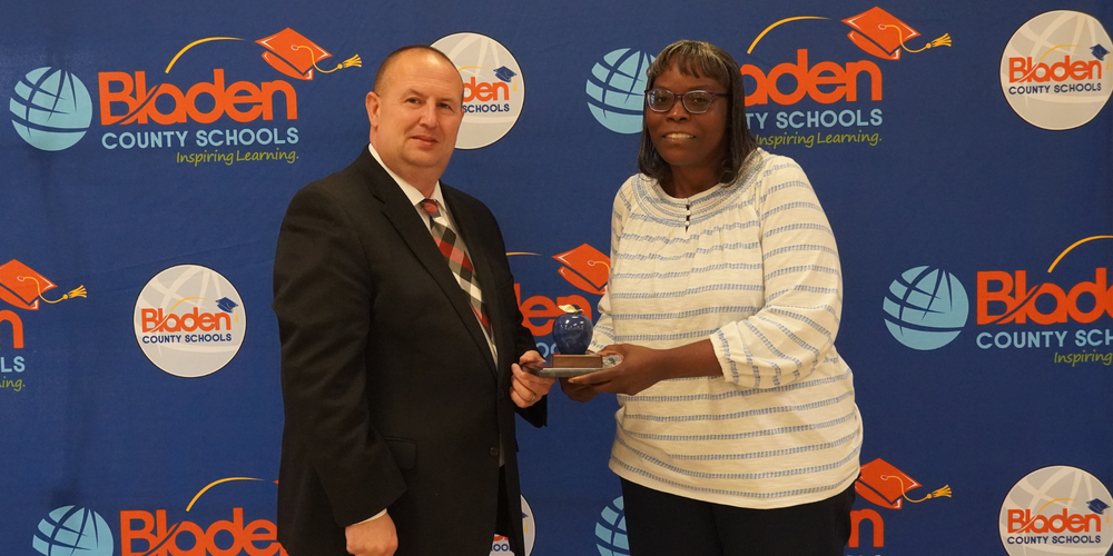 Bladen County Schools CTE Teacher of the Year stands in front of a district backdrop smiling with the superintendent.