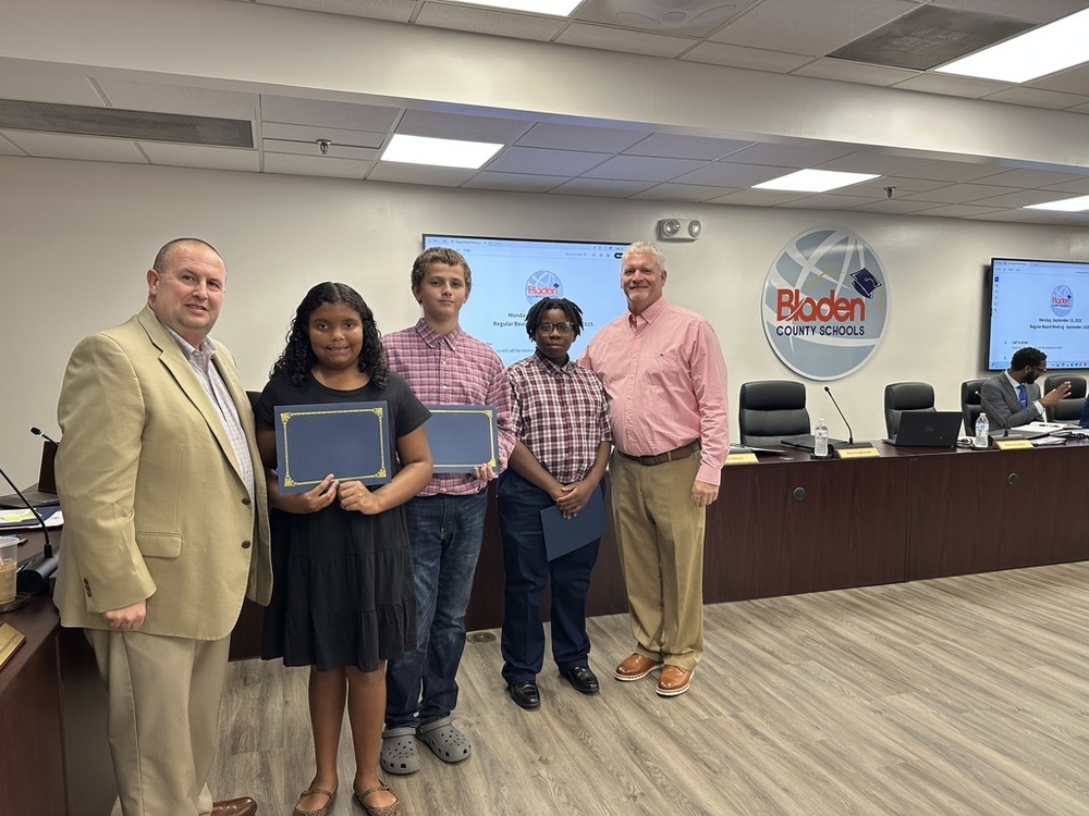 Three students stand at the front of a boardroom holding certificates, joined by two adult men in professional attire. The Bladen County Schools logo is displayed on the wall behind them.