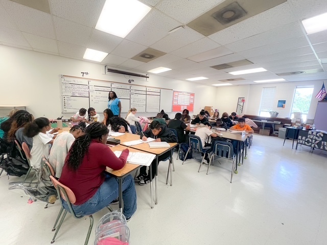 A classroom of middle school students sits in groups at desks, focused on writing assignments while a teacher stands at the back of the room.