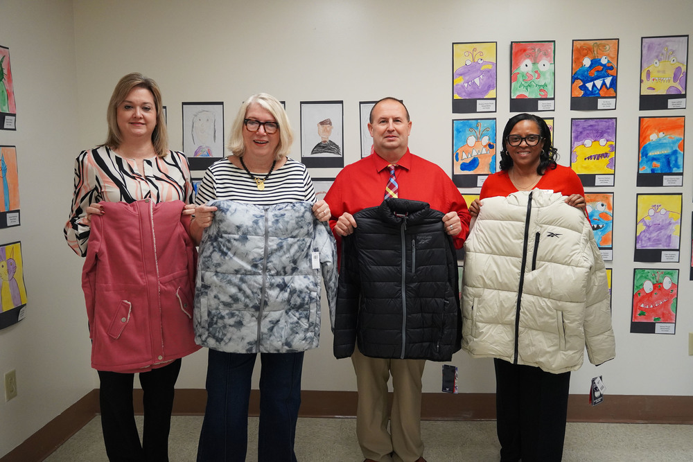 Four adults stand side by side in front of a wall decorated with colorful student artwork, each holding a winter coat. From left to right, a woman in a patterned blouse holds a pink coat; retired educator Jane Winters, wearing a striped shirt and glasses, holds a gray and white coat; a man in a red shirt and plaid tie holds a black coat; and a woman in a red top holds a cream-colored coat.