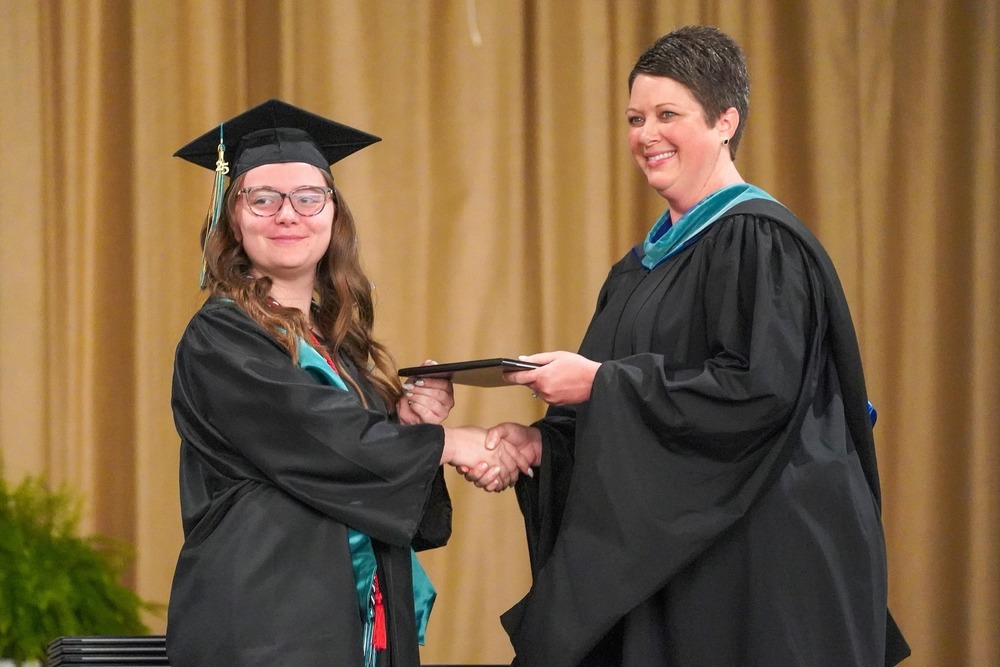 A Bladen Early College High School graduate in a black cap and gown shakes hands with a school administrator while receiving her diploma during the graduation ceremony. Both are smiling and dressed in graduation regalia, with a gold curtain backdrop behind them.
