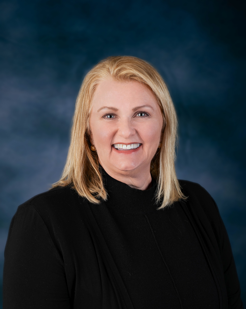 Portrait of a smiling woman with shoulder-length blonde hair, wearing a black turtleneck sweater, posed against a dark blue studio background.