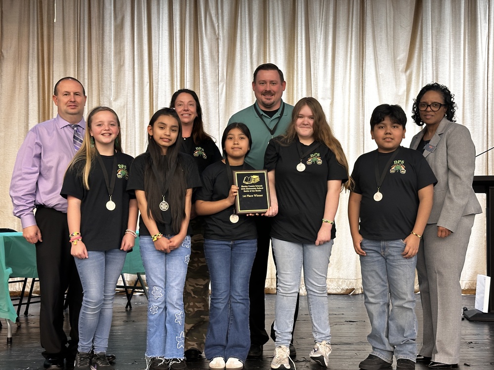 A group of elementary students wearing matching black DPS shirts and medals stand on a stage with school staff and administrators. One student holds a plaque recognizing the team as 1st Place winners in the Bladen County Schools Elementary Battle of the Books competition. A gold curtain backdrop hangs behind the group.