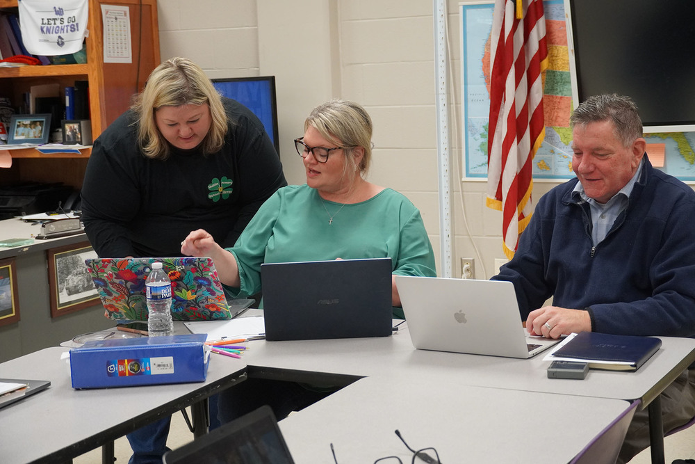 Three educators collaborate around laptops and printed data reports in a classroom, reviewing standards and instructional priorities.