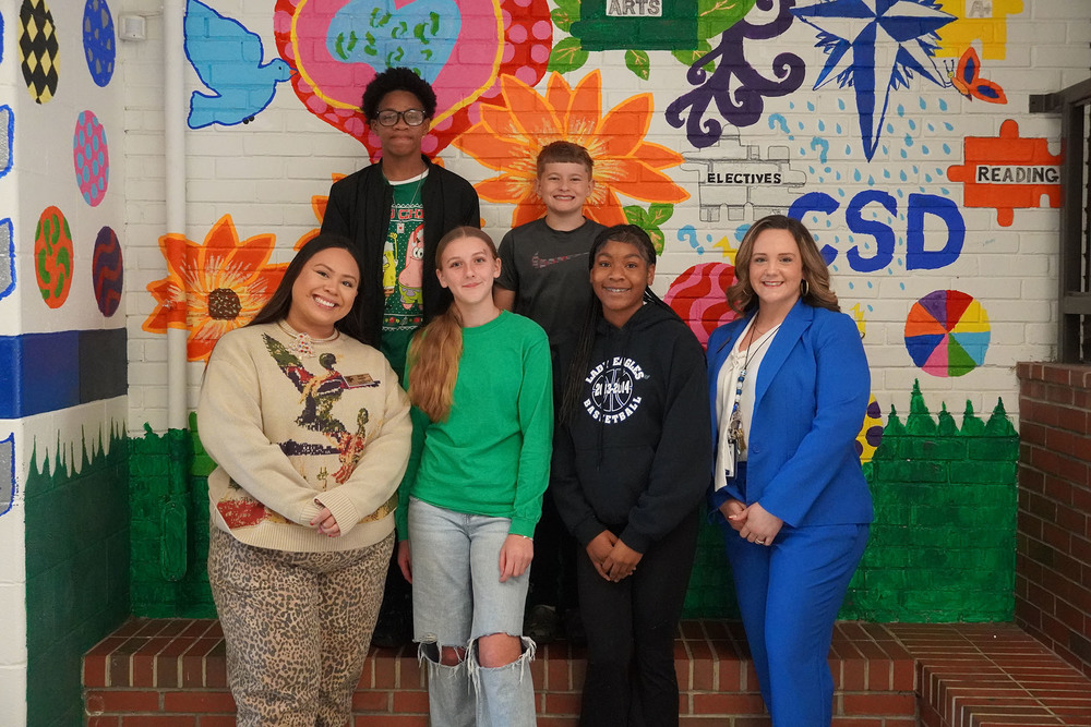 Group of students and staff pose smiling in front of a colorful school mural featuring flowers and academic subjects.
