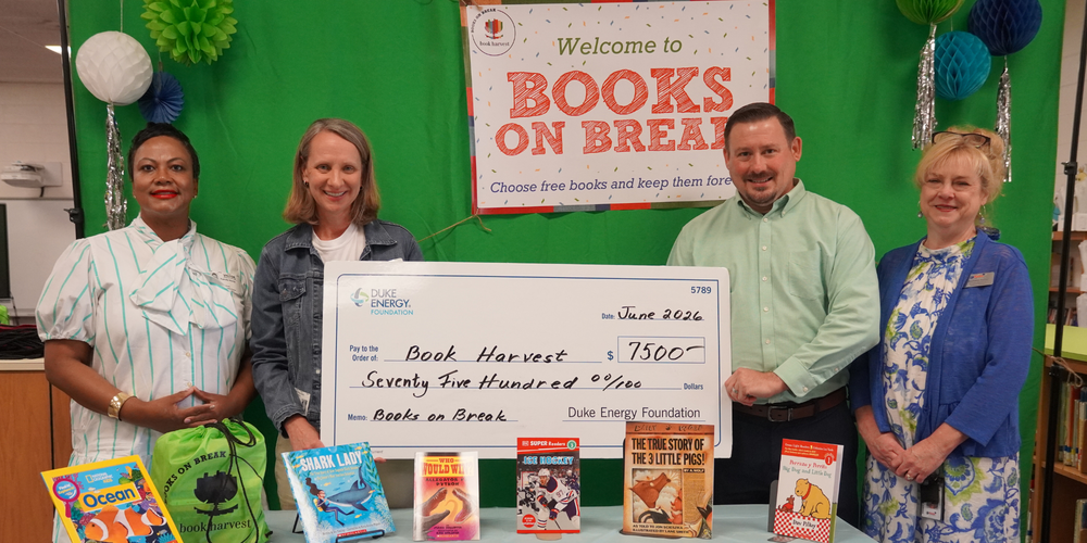 Four adults stand together holding a large check made out to Book Harvest for $7500 for Books on Break. The adults are standing behind a table with books on display.