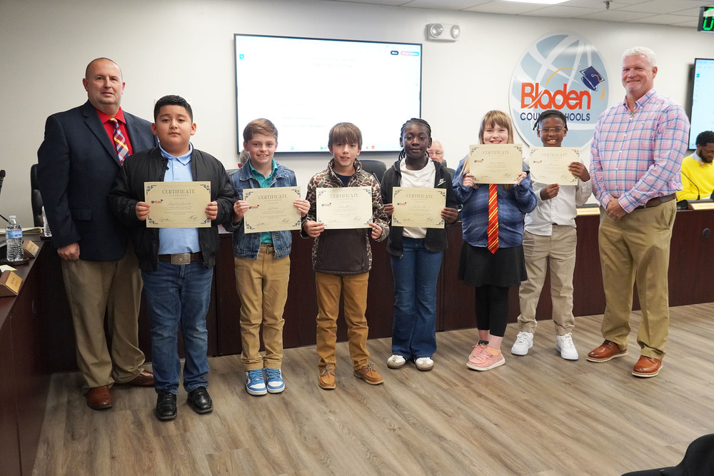 Seven elementary students stand in a row holding certificates, flanked by two adult administrators at a Bladen County Schools meeting.