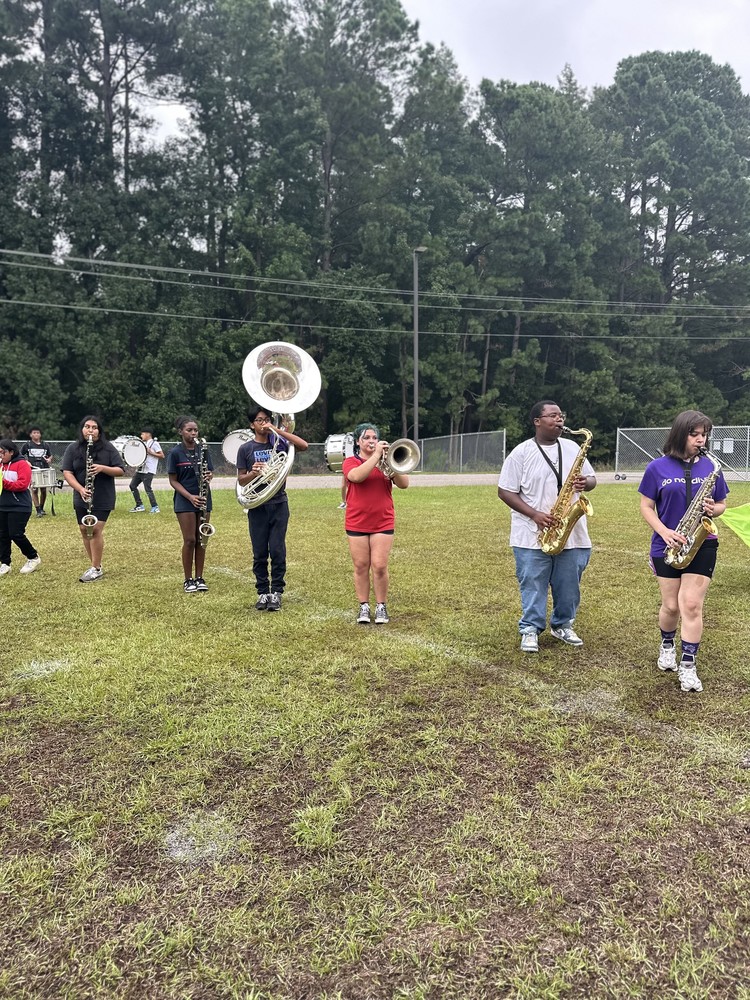 A group of high school marching band members are practicing on a grassy field surrounded by tall pine trees. The students are arranged in a line, each playing different brass and woodwind instruments, including sousaphones, saxophones, and trumpets. They are dressed in casual athletic wear, and the field has patches of muddy grass, indicating recent rain.  A chain-link fence can be seen in the background. The overall atmosphere suggests a band camp or rehearsal session. 