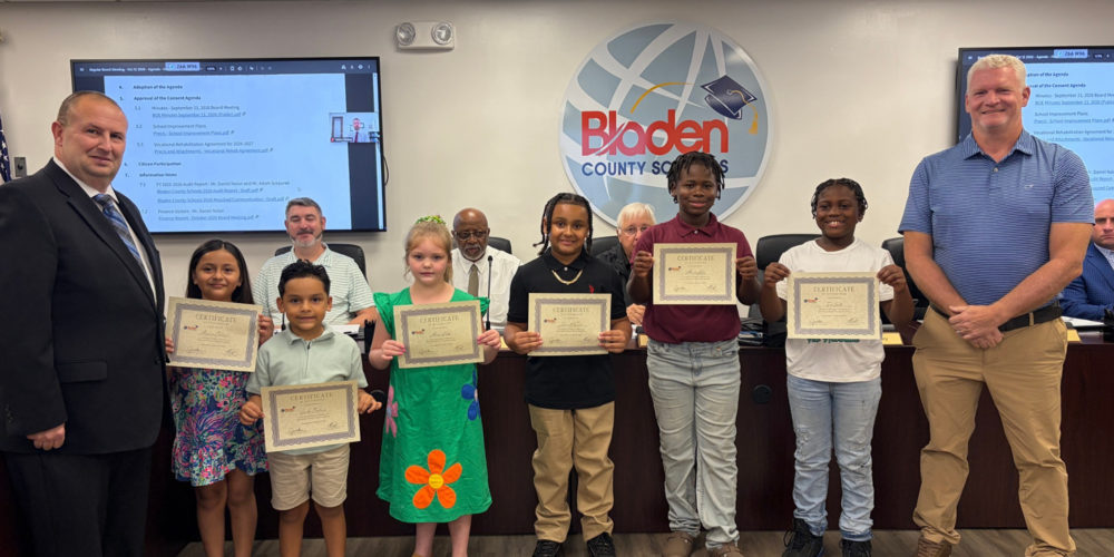 A group of elementary students holding certificates stand smiling with the superintendent and board chair.