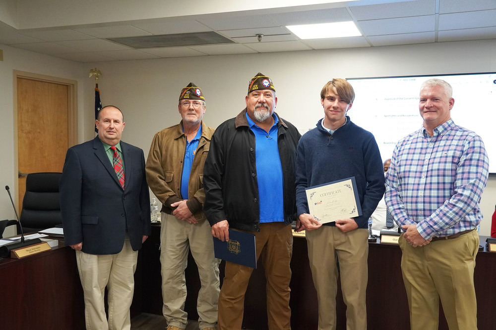 Five individuals stand in a boardroom for a recognition photo. Two veterans wearing service caps stand at the center, with a student holding a certificate beside them. A man in a suit and a man in a plaid shirt stand on either side, all facing the camera.