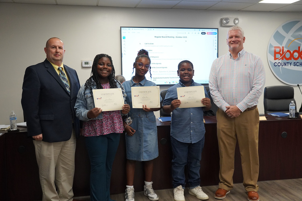 Three elementary students stand in the center of a boardroom holding certificates of recognition, smiling proudly between two adult men in professional attire. Behind them, a presentation slide on the wall displays the October 2025 Regular Board Meeting agenda and a large Bladen County Schools logo.