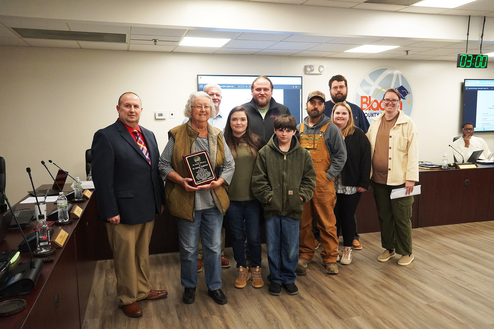 A group of family members stands with an older woman holding a commemorative plaque during a Bladen County Schools Board of Education meeting. District leaders stand alongside them in the boardroom.