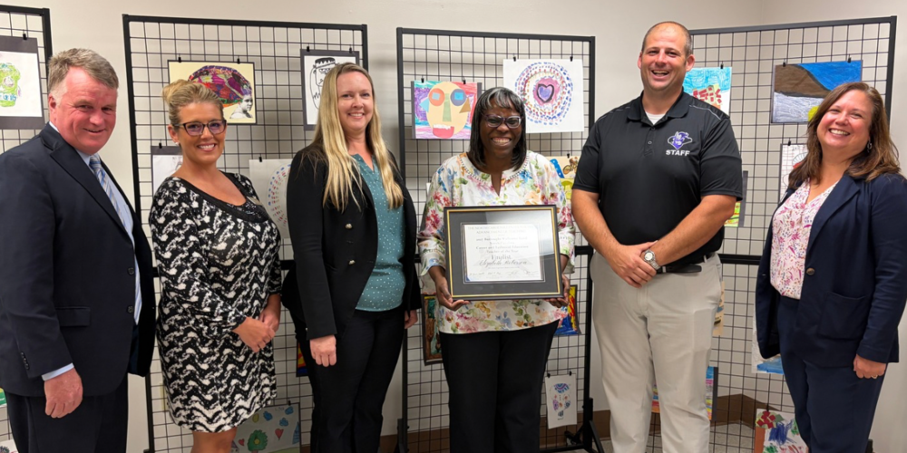 Six adults stand smiling together in front of student art. One individual is holding a framed certificate.