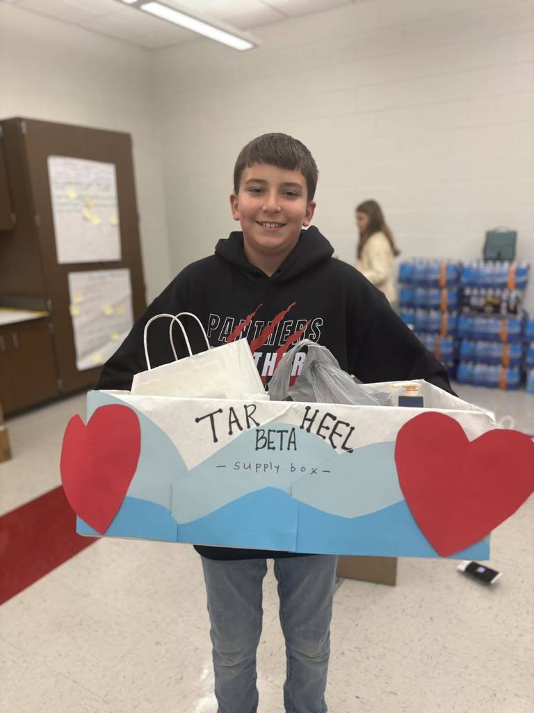 A student smiles while holding a decorated box labeled "Tar Heel Beta Supply Box," filled with donated supplies. Large red hearts decorate the sides of the box.