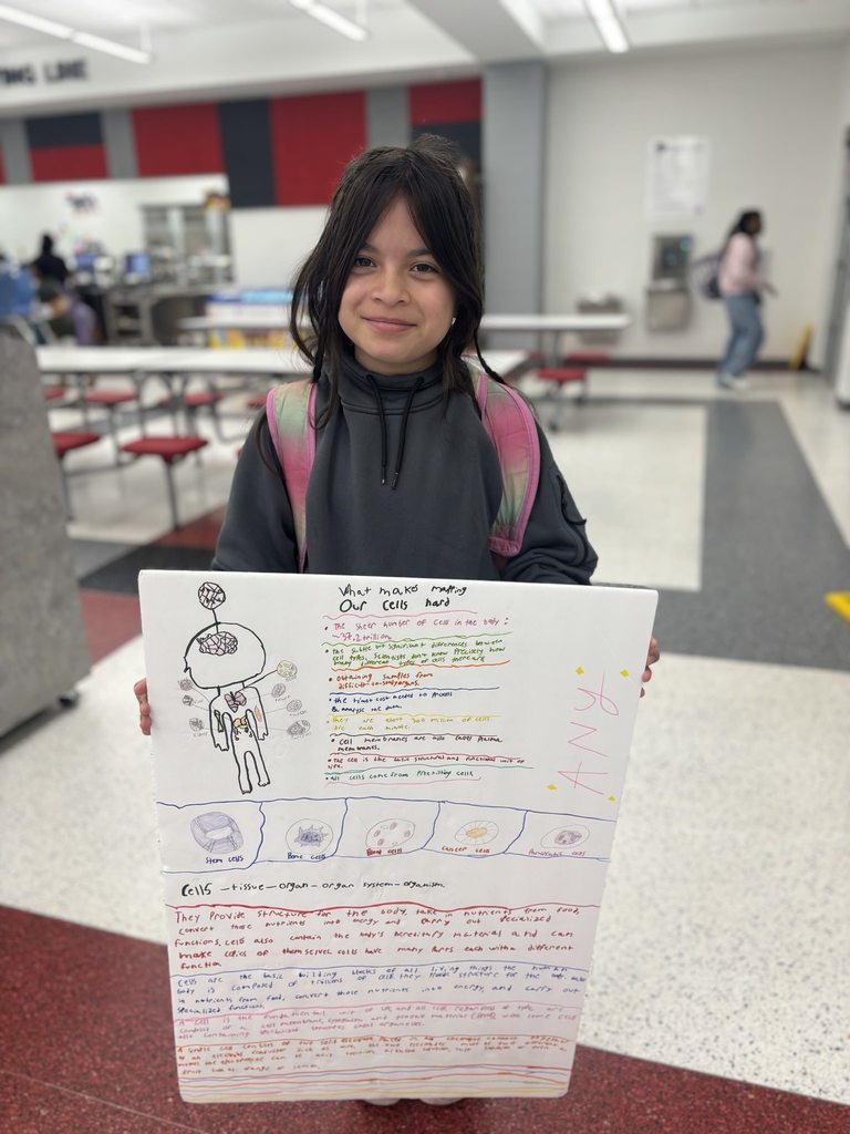 A student smiles while holding a large poster about cells, featuring hand-drawn illustrations and colorful handwritten notes, in a school cafeteria.