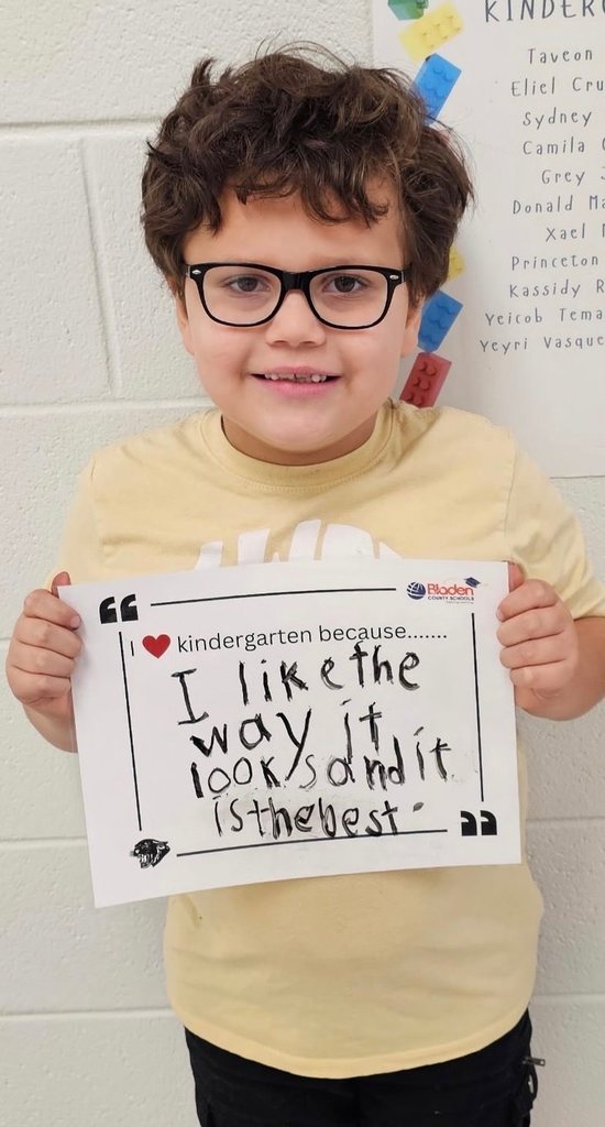A kindergarten student smiles while holding a sign that reads, “I ❤️ kindergarten because… I like the way it looks and it is the best.”