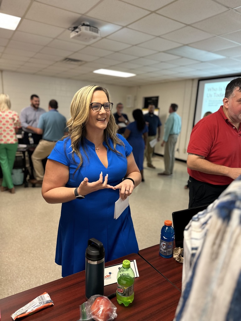 A school leader in a bright blue dress speaks with enthusiasm during a leadership meeting, holding a notecard and gesturing while engaged in conversation. The table in front of her displays a nameplate, snacks, and drinks, while colleagues collaborate in the background.