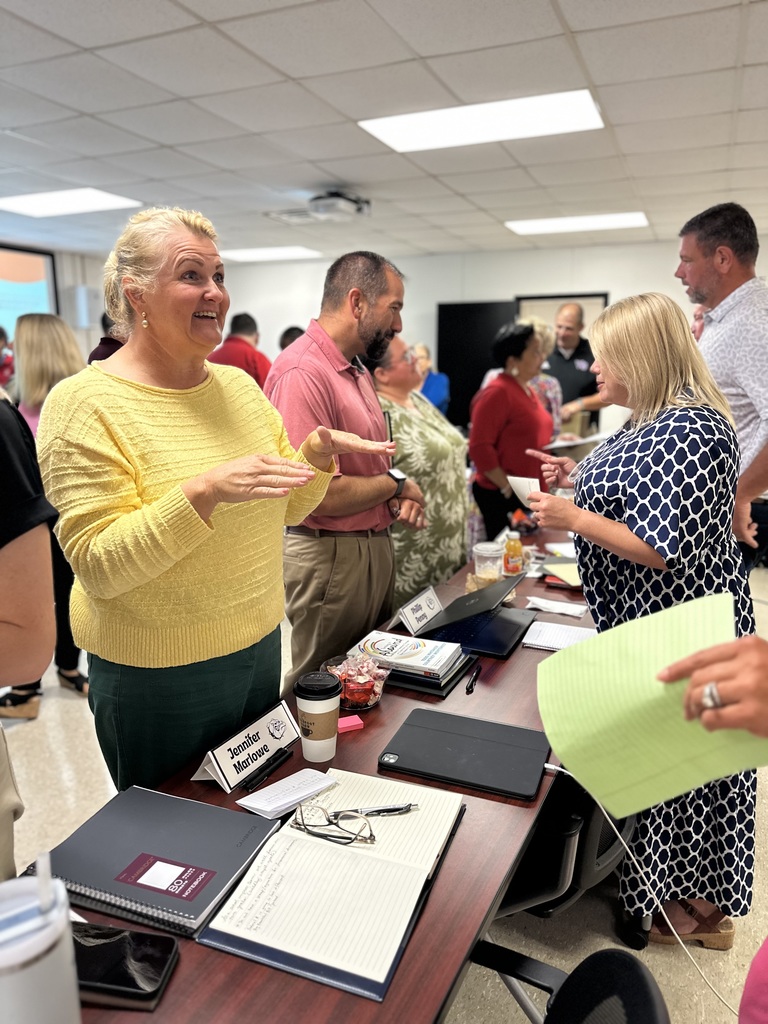 Leaders engage in animated conversation during a professional development session. A woman in a yellow sweater gestures enthusiastically across the table, while others listen and contribute in the background. The table is filled with notebooks, laptops, coffee, and name tags, reflecting active collaboration and planning.