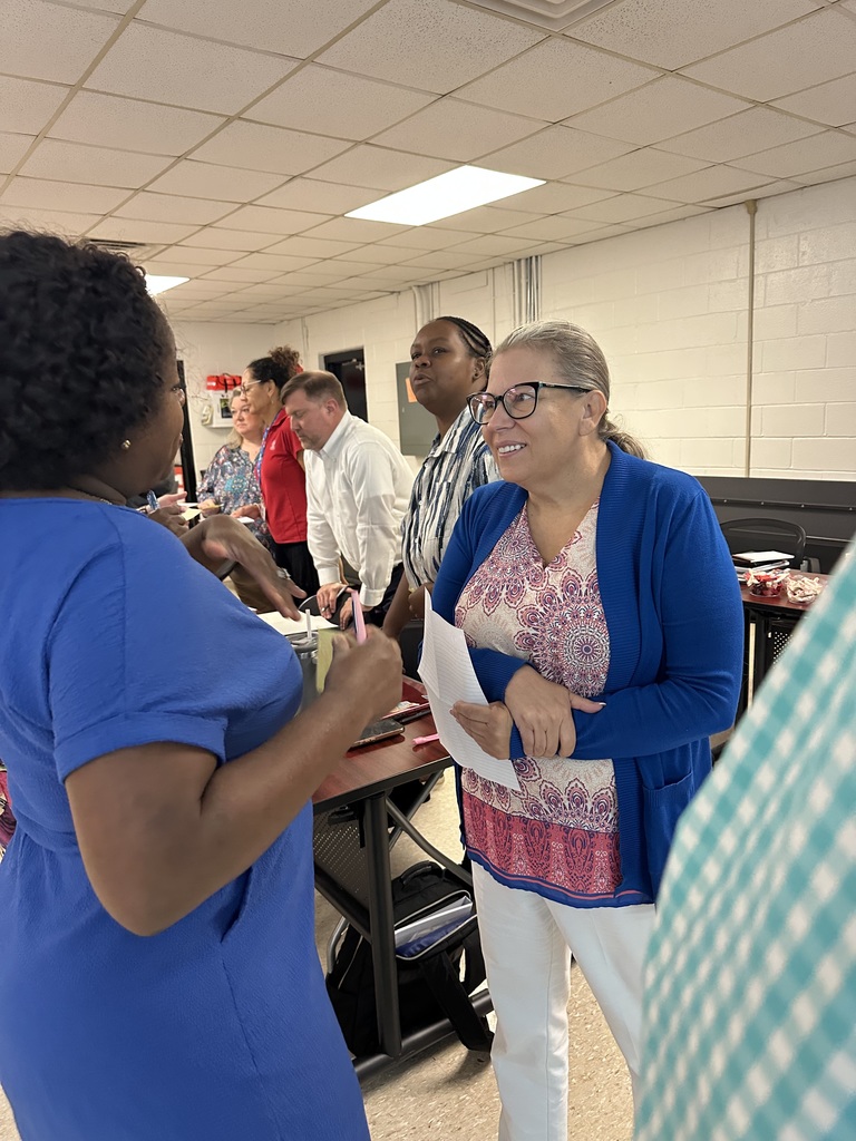 School and district leaders engage in conversation during a leadership meeting. In the foreground, two women share ideas with smiles and notes in hand, while other team members collaborate in the background, reflecting a positive and collaborative atmosphere.