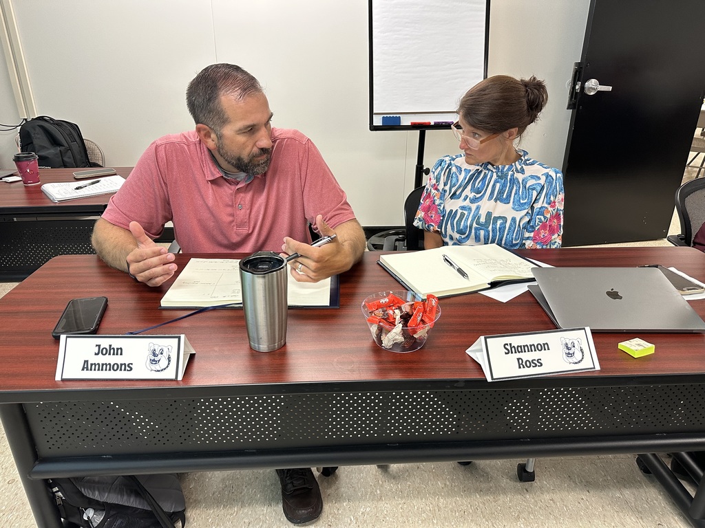 Two school leaders, seated at a table labeled with nameplates for John Ammons and Shannon Ross, engage in focused discussion during a leadership meeting. Notebooks, a laptop, and a bowl of candy are spread out on the table, reflecting active collaboration and planning.