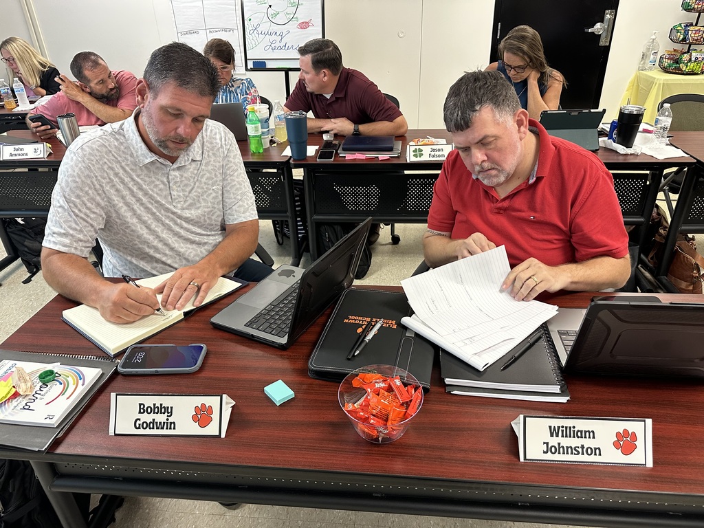 School leaders Bobby Godwin and William Johnston focus on their work during a leadership session. Seated at a shared table with nameplates, laptops, notebooks, and materials, they review notes and write as other team members collaborate in the background, reflecting dedication to planning for the year ahead.