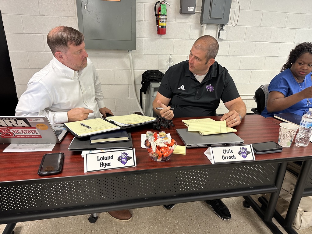 Leland Hyer and Chris Orrock sit side by side at a table during a leadership meeting, engaged in discussion with notebooks, laptops, and nameplates in front of them. A bowl of candy and bottled water suggest a productive and comfortable working environment. A third participant is seated to the right, writing on a notepad.