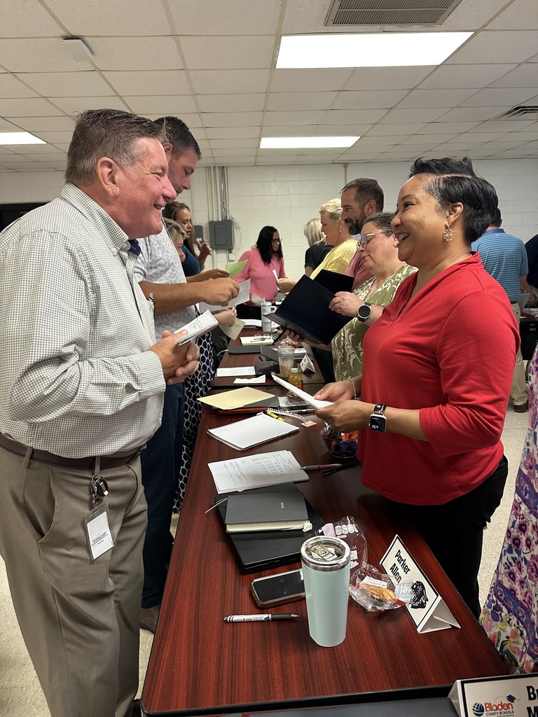 School leaders participate in a table activity during a leadership meeting, exchanging ideas and documents with enthusiasm. A man and woman smile as they engage in discussion, surrounded by notebooks, nameplates, and snacks, highlighting a collaborative and energized atmosphere.