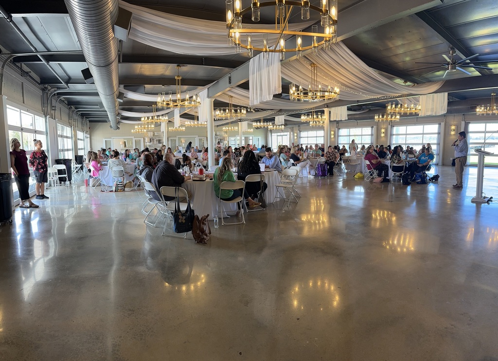Large group of staff seated at round tables inside a decorated event space listening to a speaker.