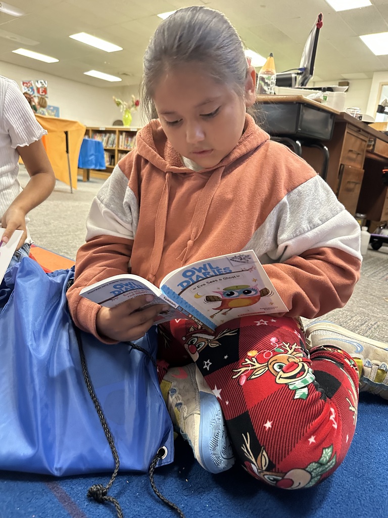 A young student sits on the classroom floor reading a book titled Owl Diaries: Eva Sees a Ghost. She is wearing a rust-colored hoodie and red holiday-themed pants with reindeer designs, with a blue drawstring bag resting beside her. The setting appears to be a school library or classroom with bookshelves and desks in the background.